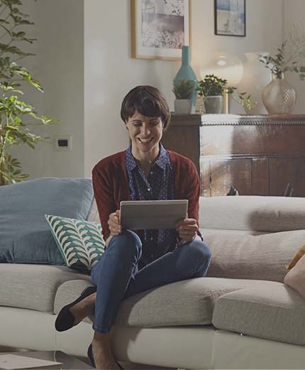 Scatto di giovane donna sorridente comodamente seduta sul divano di casa che apre il conto da tablet