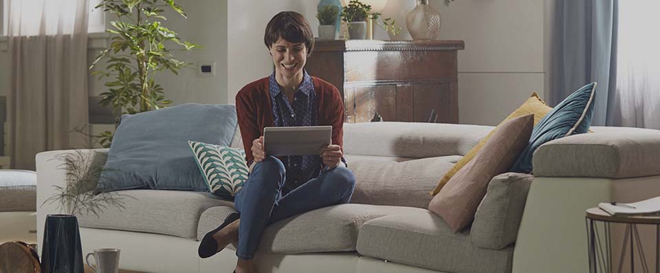 Scatto di giovane donna sorridente comodamente seduta sul divano di casa che apre il conto da tablet