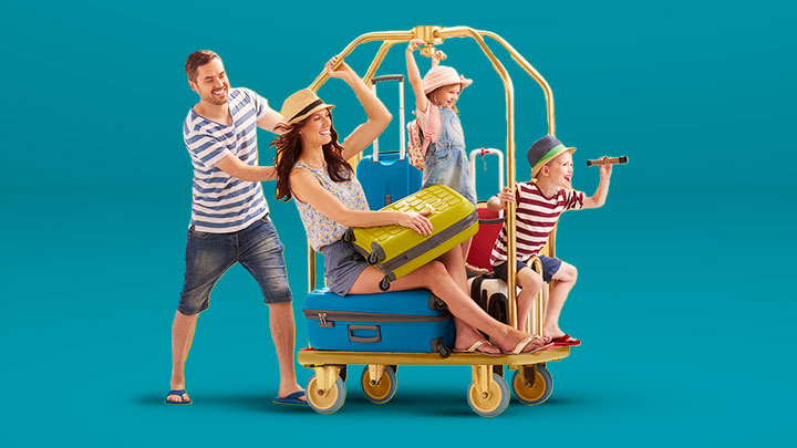 Giovane papà spinge per gioco un carrello bagagli con sopra mamma, figlio e figlia. Tutti indossano abiti molto sportivi e colorati, la famiglia è pronta per iniziare la vacanza 