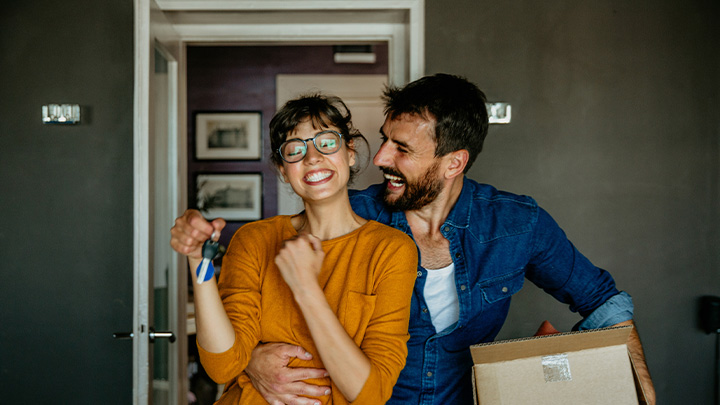 Immagine fotografica di una giovane coppia in un interno: la donna sorride e tiene in mano le chiavi di casa, l'uomo la abbraccia ridendo mentre porta uno scatolone